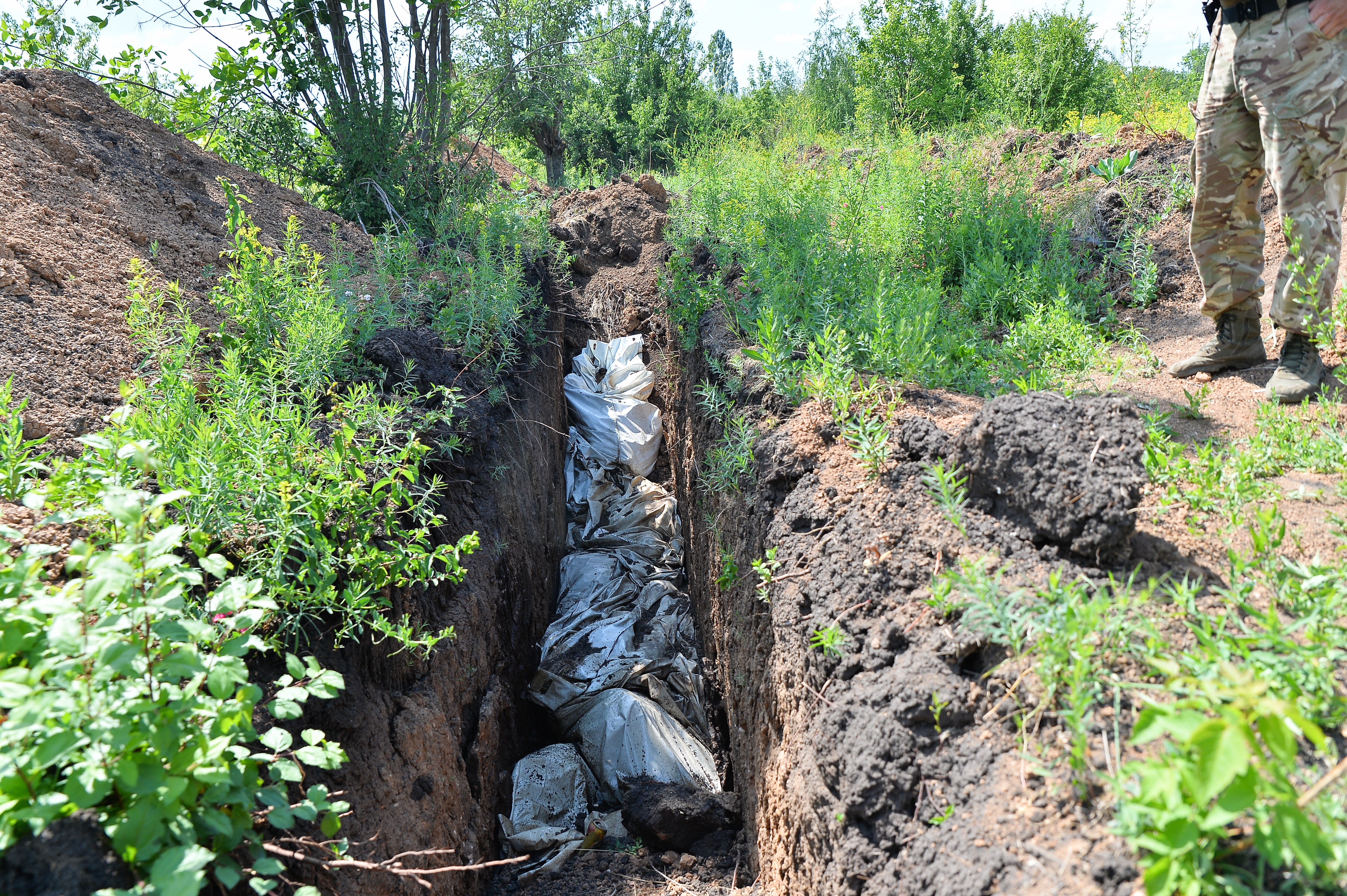 A trench in the ground shows stuffed white garbage bags lined up. One person is shown from the waist down observing the bags and the trench.