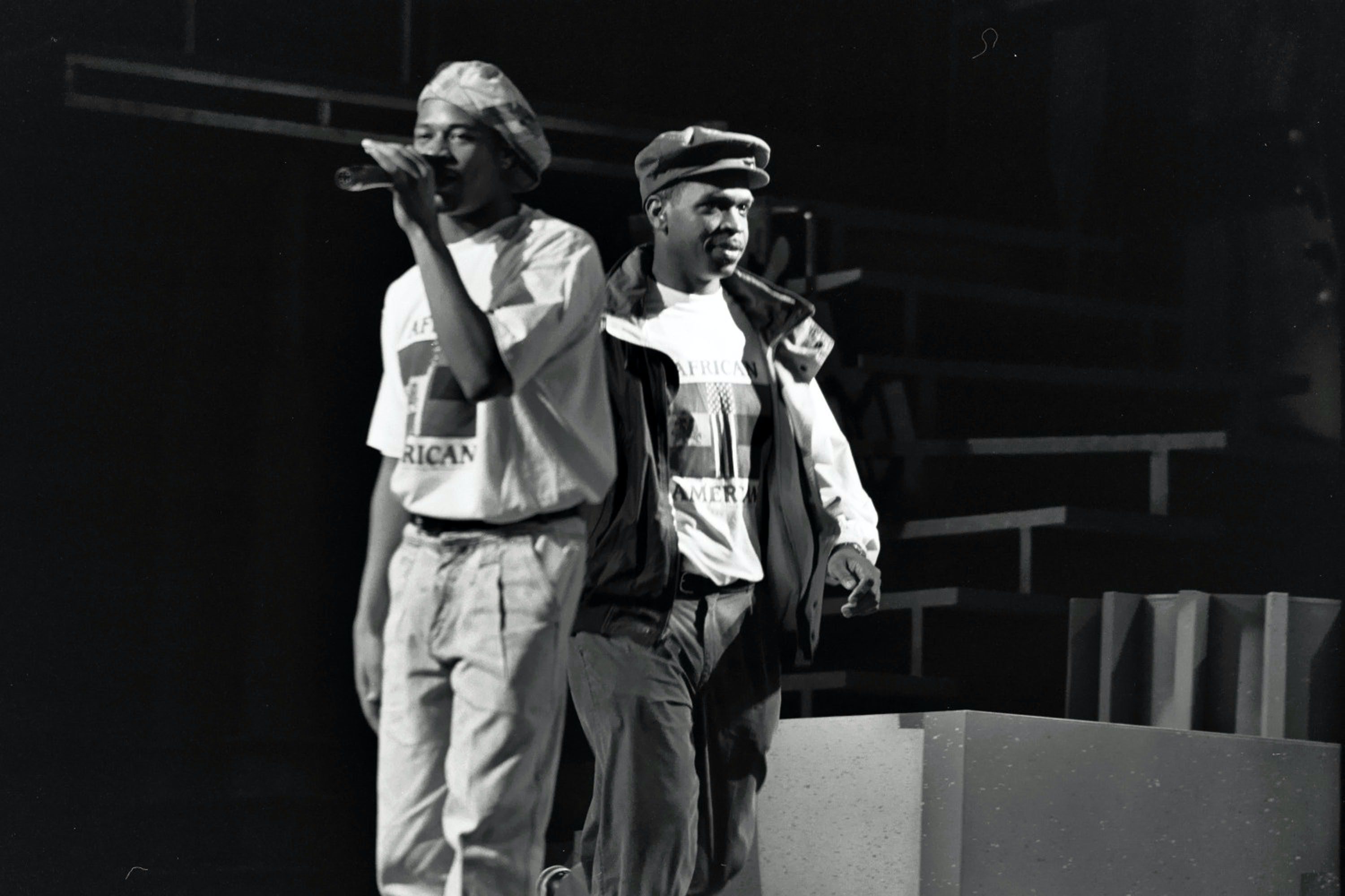 Two young Black men wearing hats stand back to back on a stage.