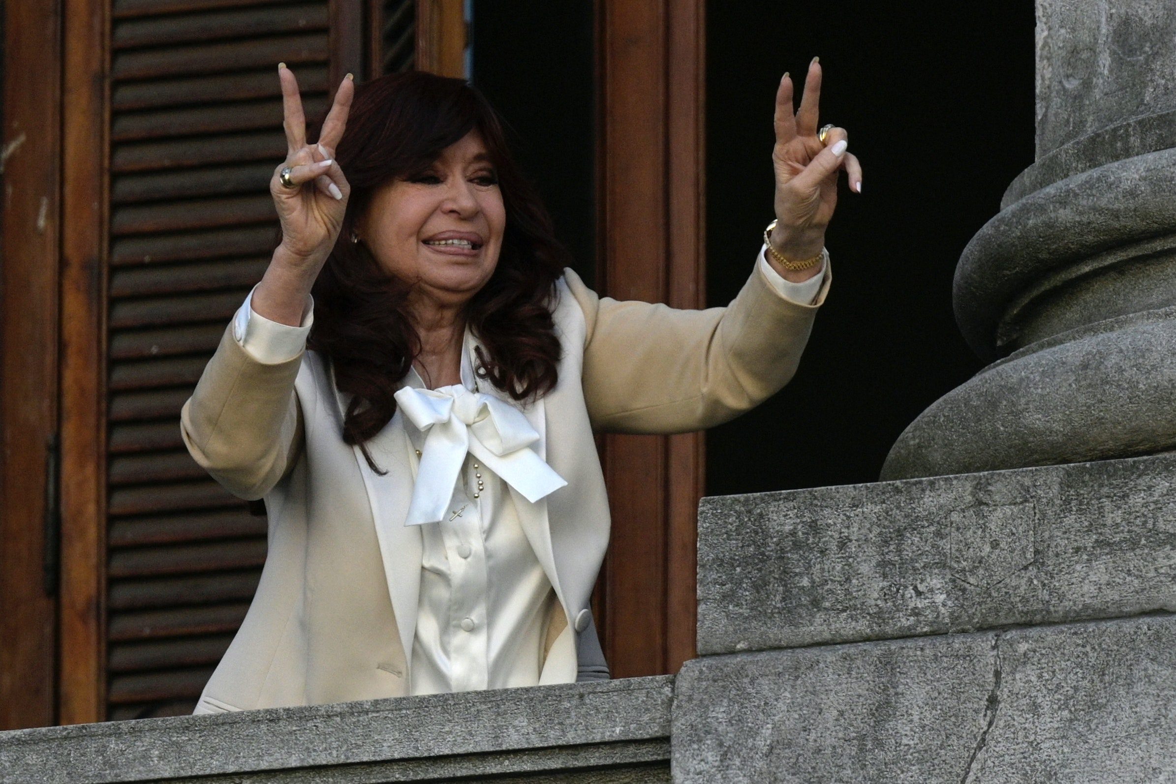 A woman wearing a beige jacket flashes a 'v for victory' sign while standing on a balcony.