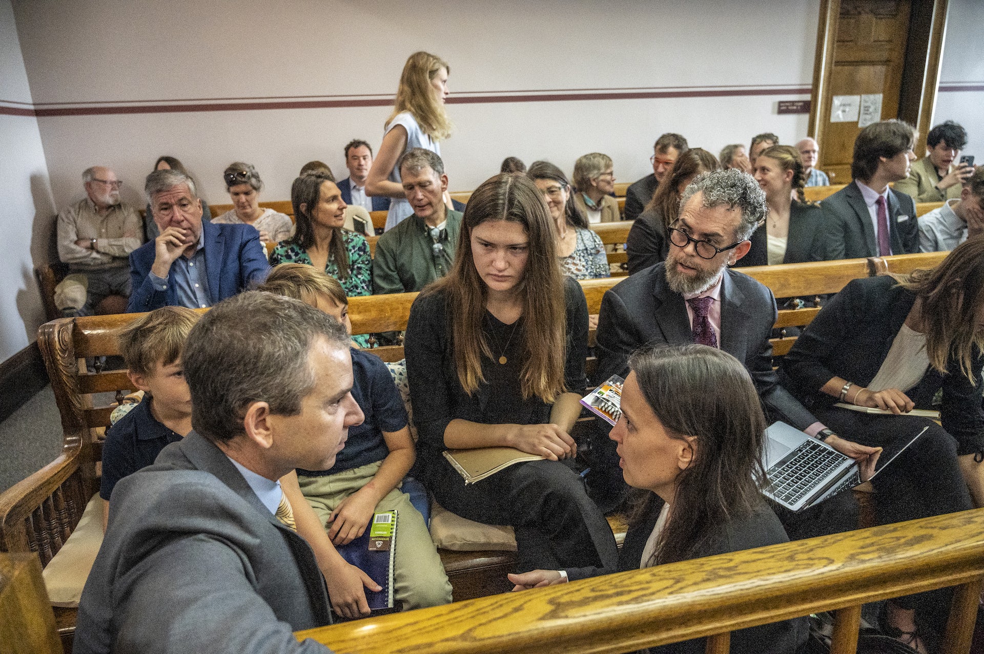 A young woman and two young boys listen as lawyers talk. Young people fill two rows of benches behind them in the small court room.