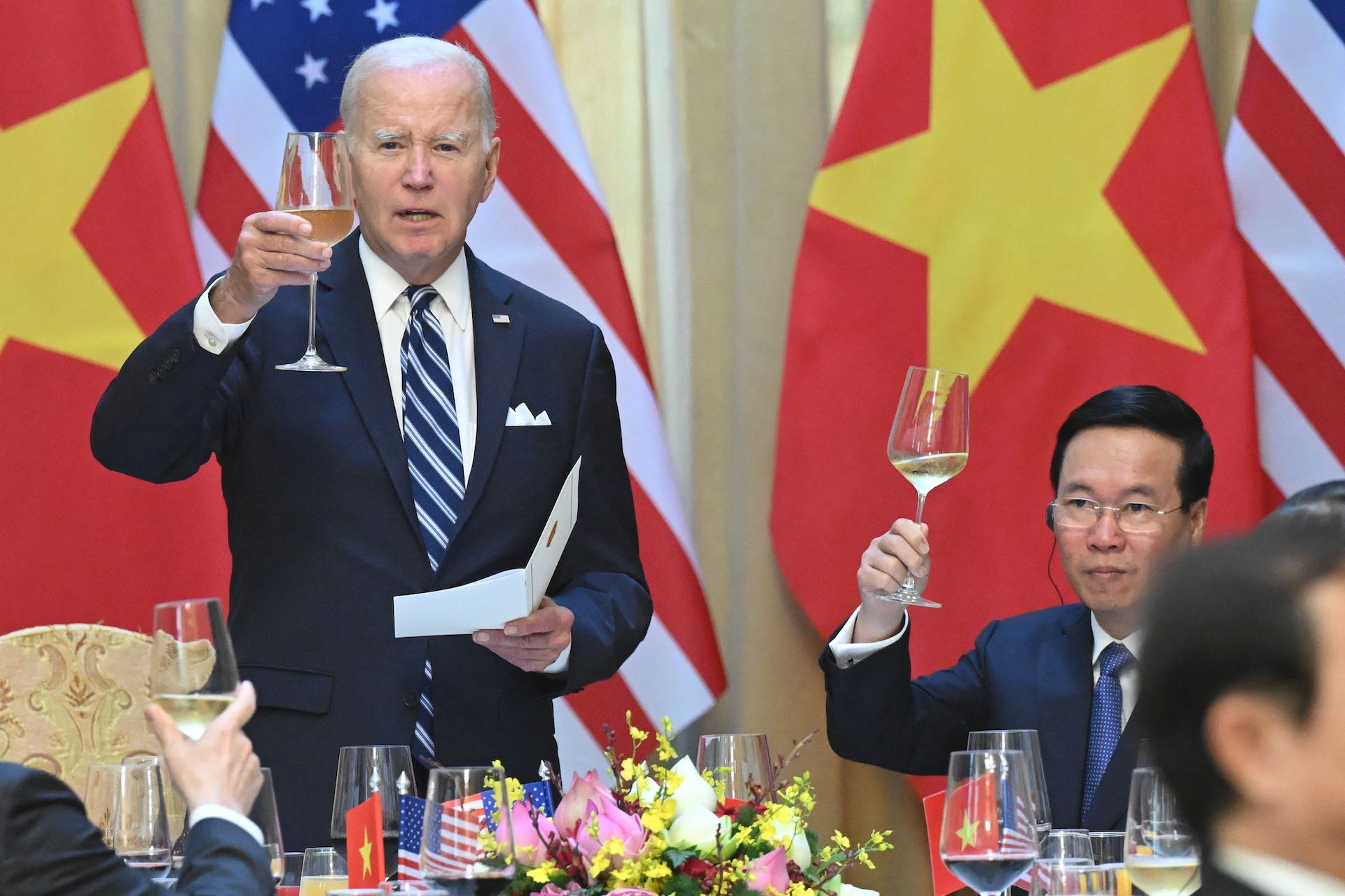 A gray haired man holds paper in his left hand as he raises a half-filled champagne glass in his right hand. To his left, a seated man wearing glasses raises his half-filled champagne glass.