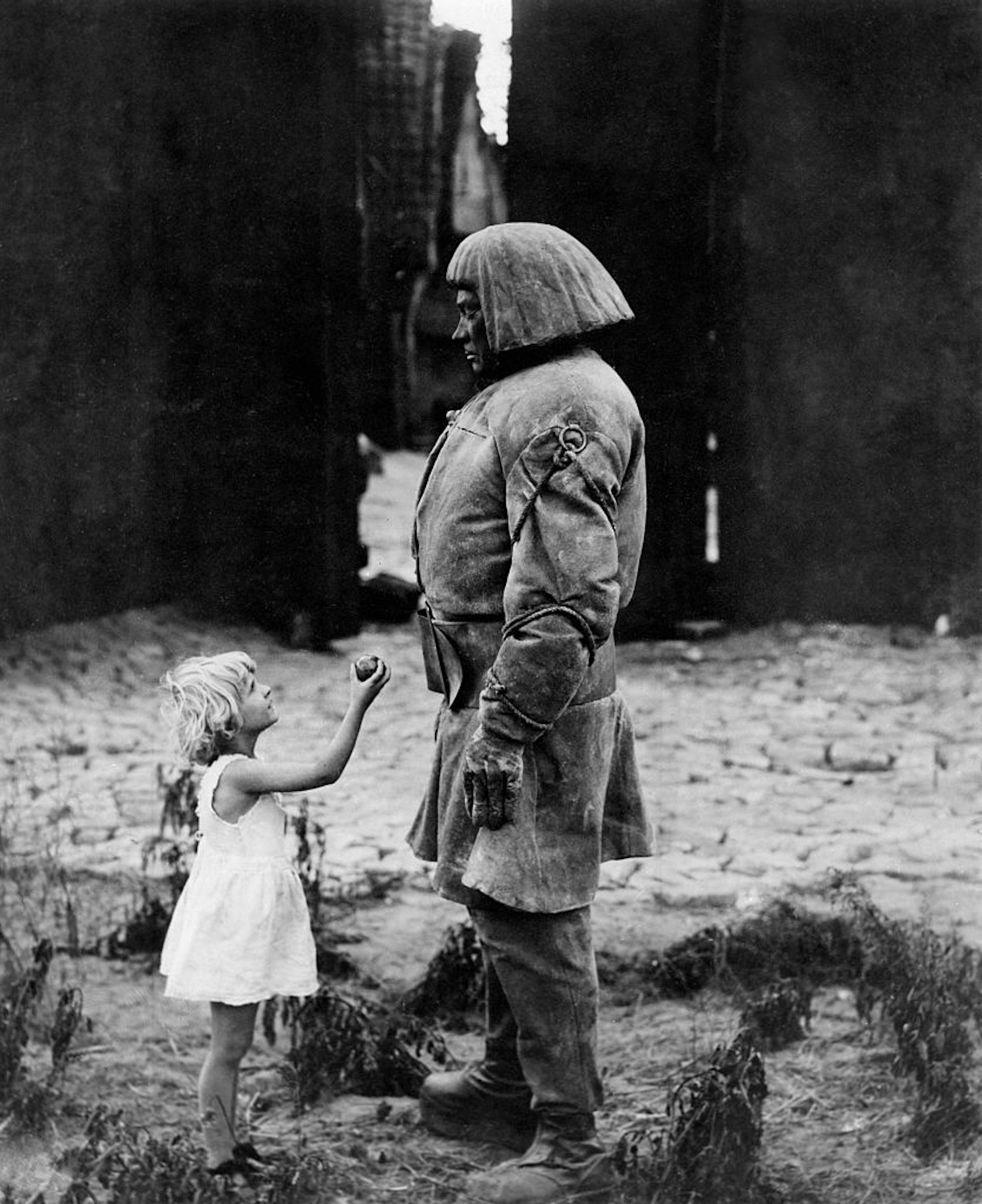 A black and white photo of a little girl in a white dress holding up a piece of fruit to a huge man in dirty clothes in an alleyway.