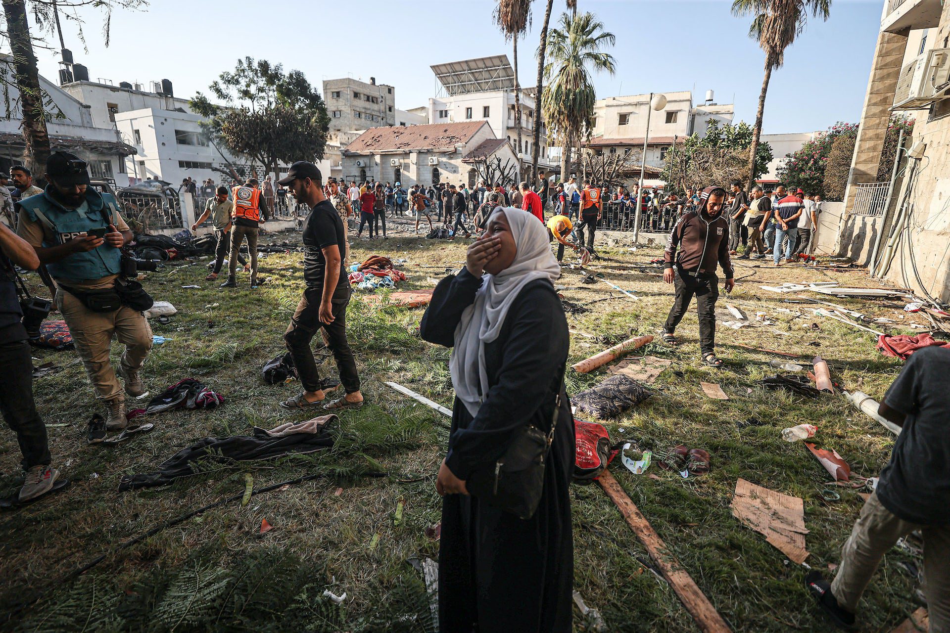 A woman holds her hand over her face and cries in a grassy field that is cluttered with people's belongings. Other people walk nearby.