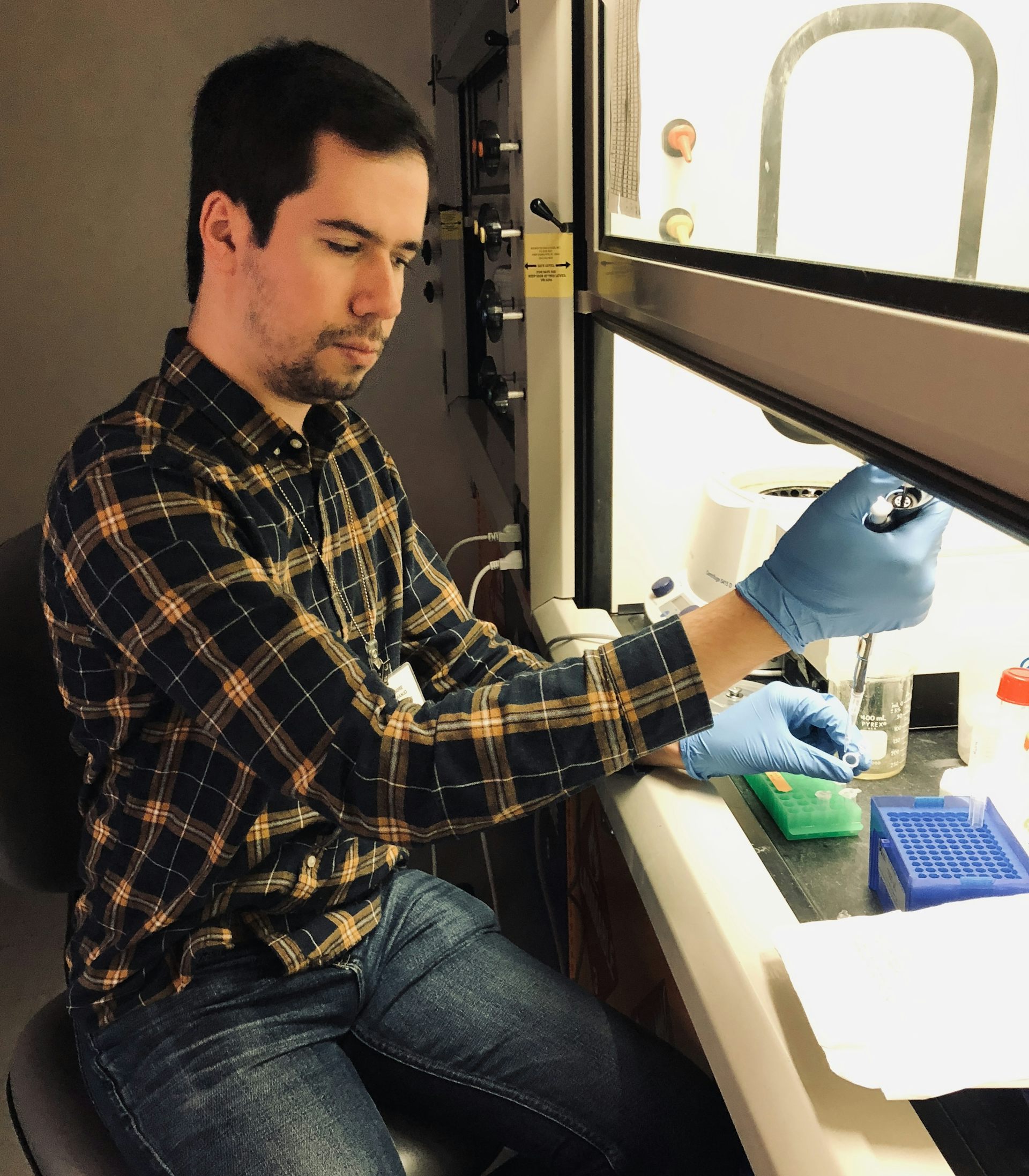 A man wearing a plaid shirt and blue rubber gloves working under a hood