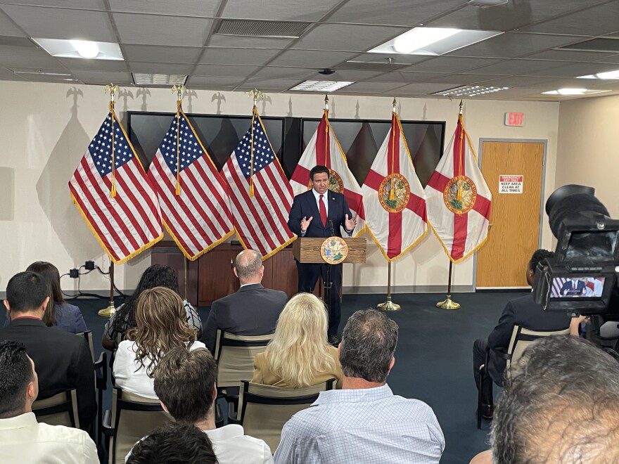 A man in a suit stands at a podium flanked by American flags and the flag of the State of Florida.