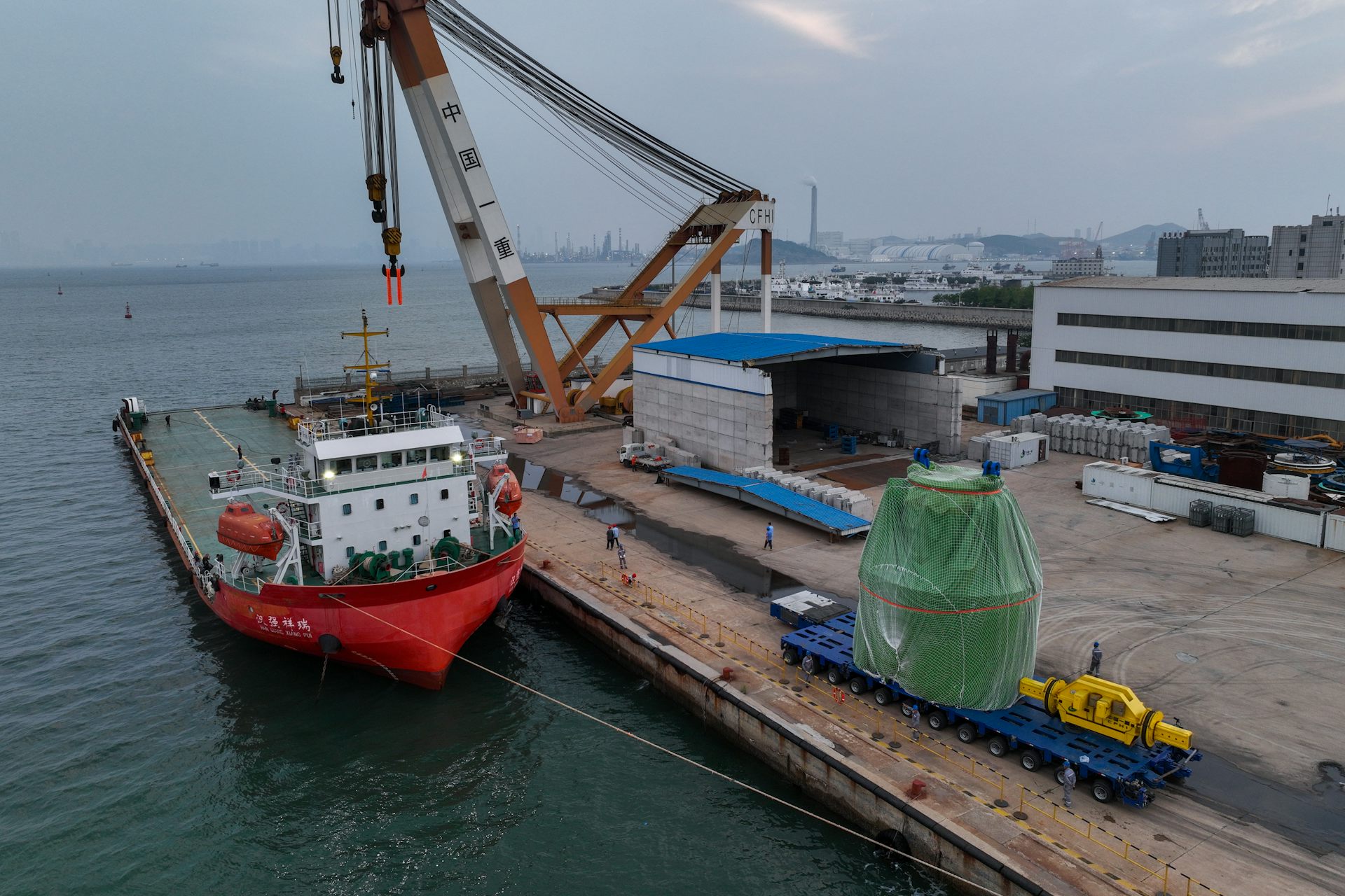 A large green item is on a pier next to a ship and a crane.