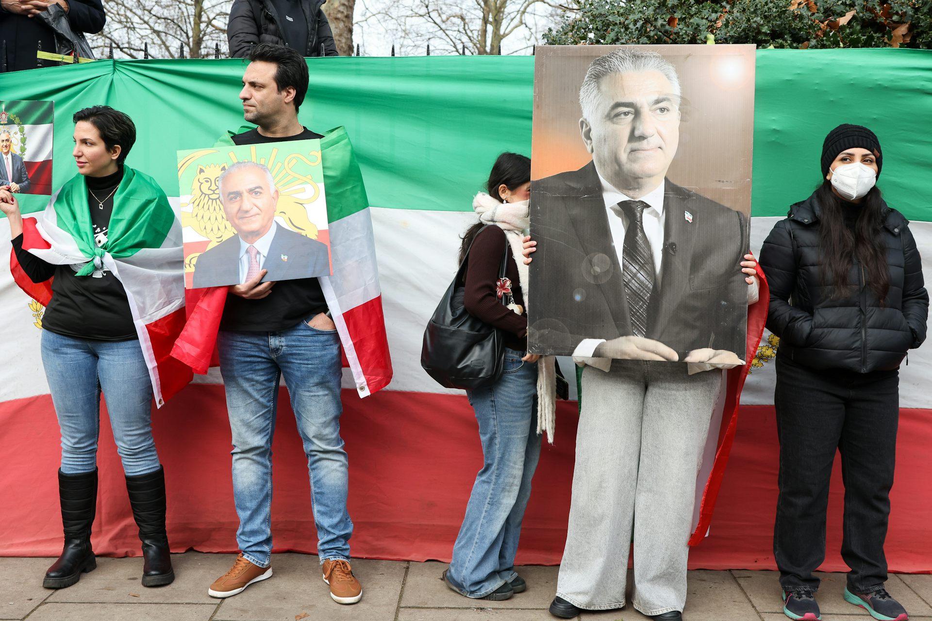 Protesters holding enlarged photos of Reza Pahlavi as they stand on a street, some of them wearing flags around their shoulders.