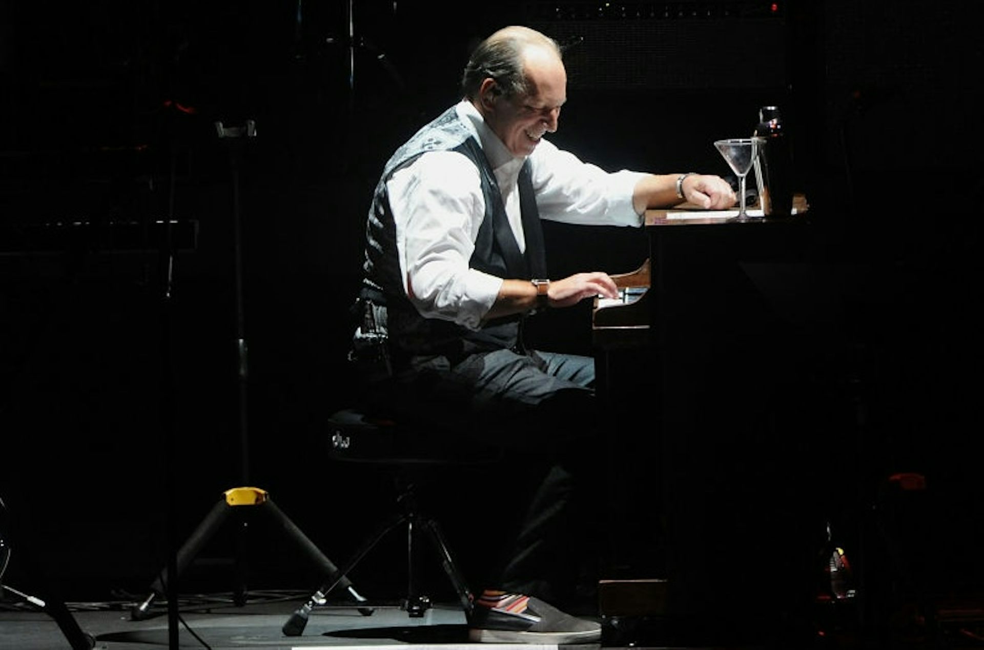 Balding, middle-aged man in a white shirt and black vest smiles while sitting at a piano on a stage.