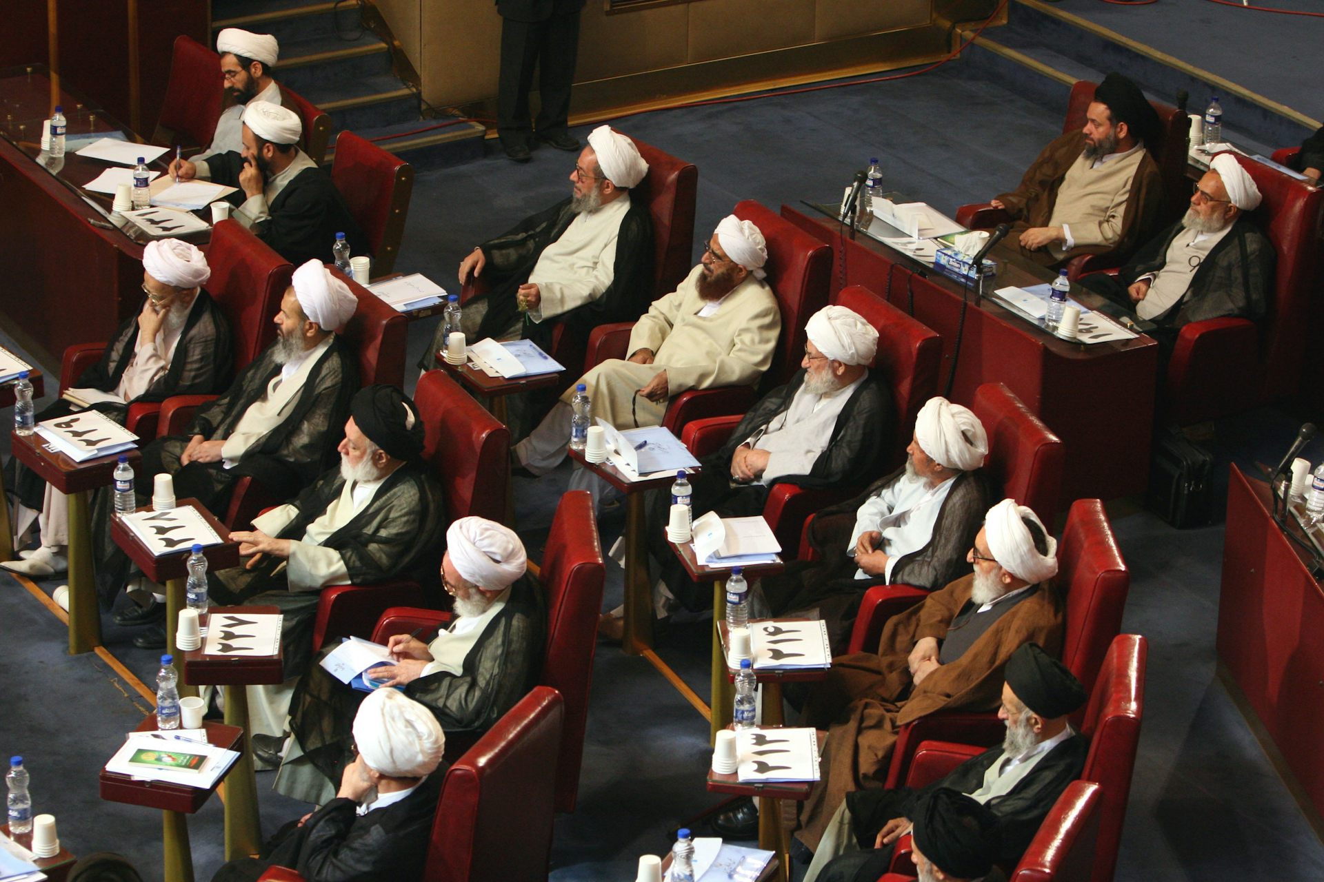 A group of clerics wearing traditional head coverings and long beards sit on seats arranged in concentric rows.